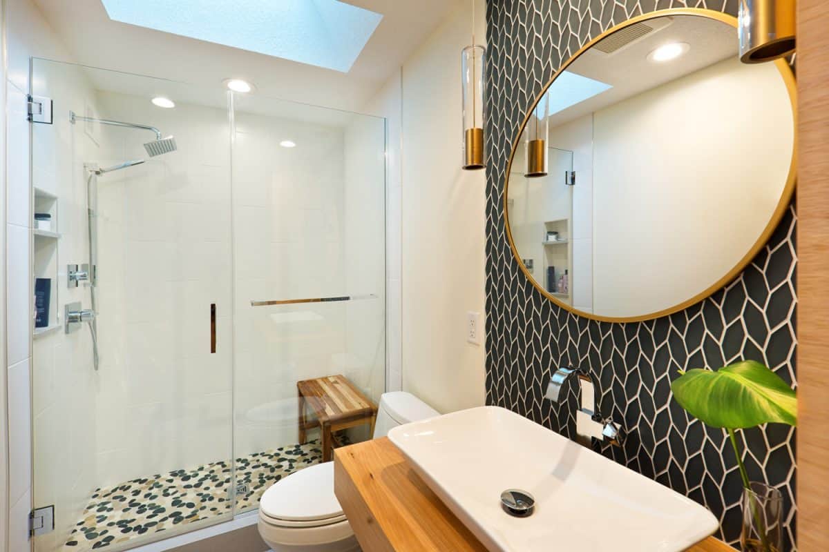 A contemporary modern bathroom design. featuring a round mirror, an above counter vessel sink, custom built vanity with storage, toilet and walk-in glass shower stall.

