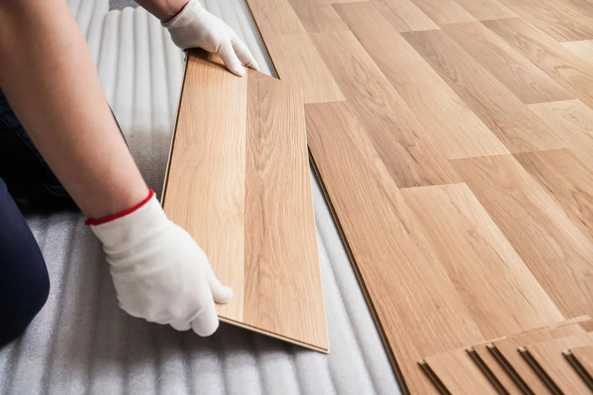 man wearing white safety gloves installing light colored wood floor tiles
