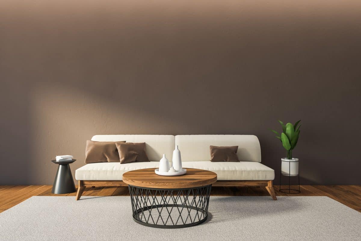 Mockup blank brown wall above sofa in living room interior with parquet floor and grey carpet. Coffee table with dishes on a plate, plant in the pot