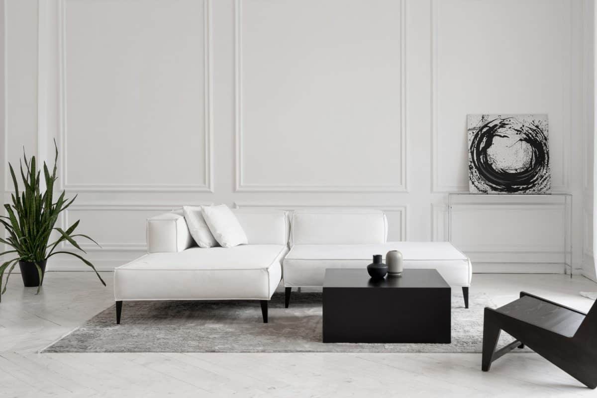 Modern white designer sofa on legs with cushions on grey carpet in middle of minimalistic living room with high ceiling, futuristic chair, green plant, abstract picture and two vases on table
