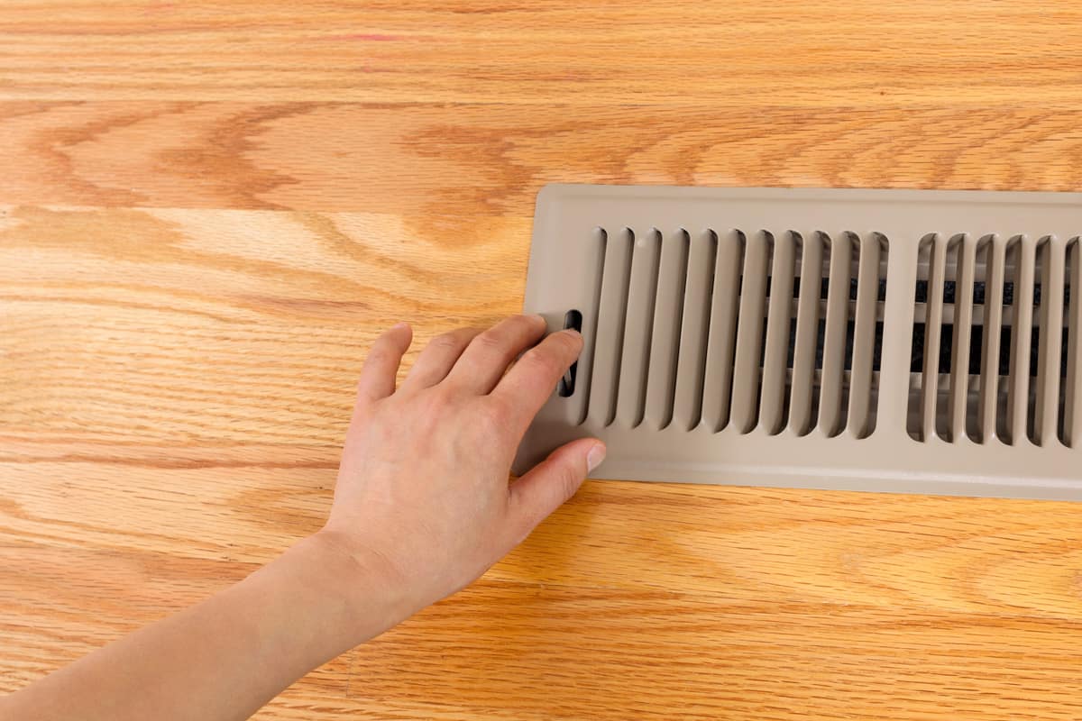 Horizontal photo of female hand opening up heater floor vent with Red Oak Floors in background.